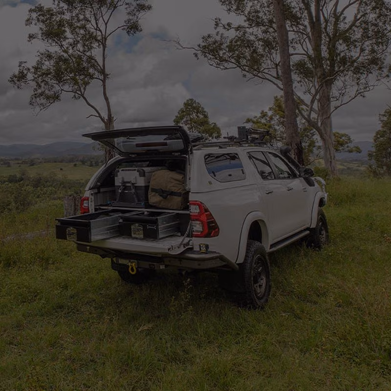 White pickup truck with a canopy, open rear drawers and a cooler, parked on a grassy hill.