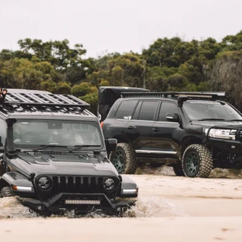 Two black off-road SUVs with roof racks and rugged tyres driving through shallow water on a sandy beach.