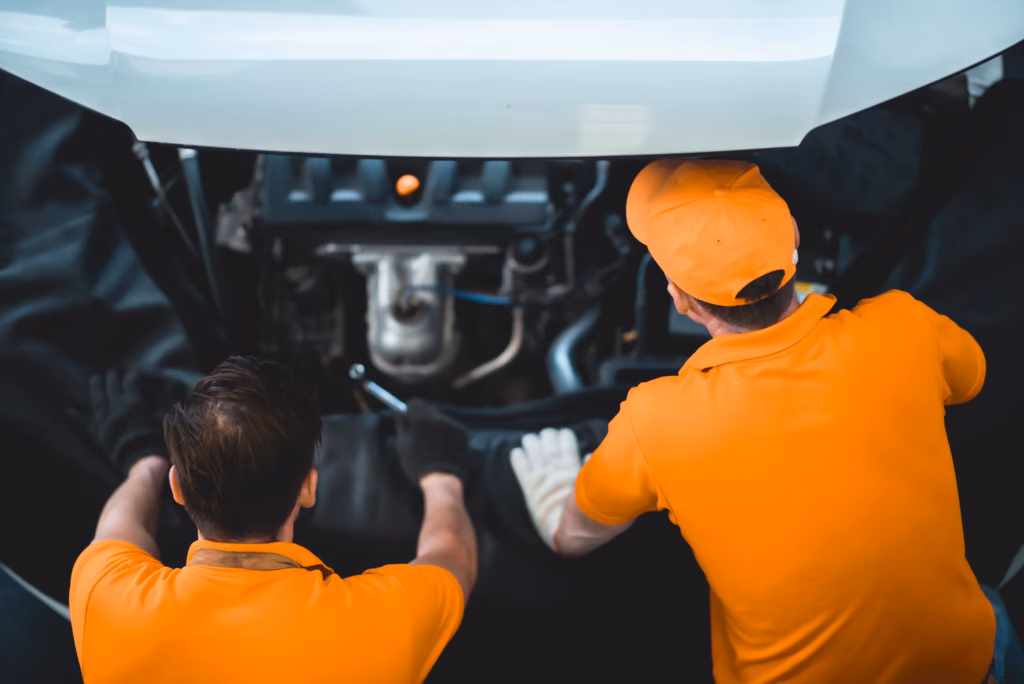 Two mechanics in orange shirts work together on a car engine with the hood raised.