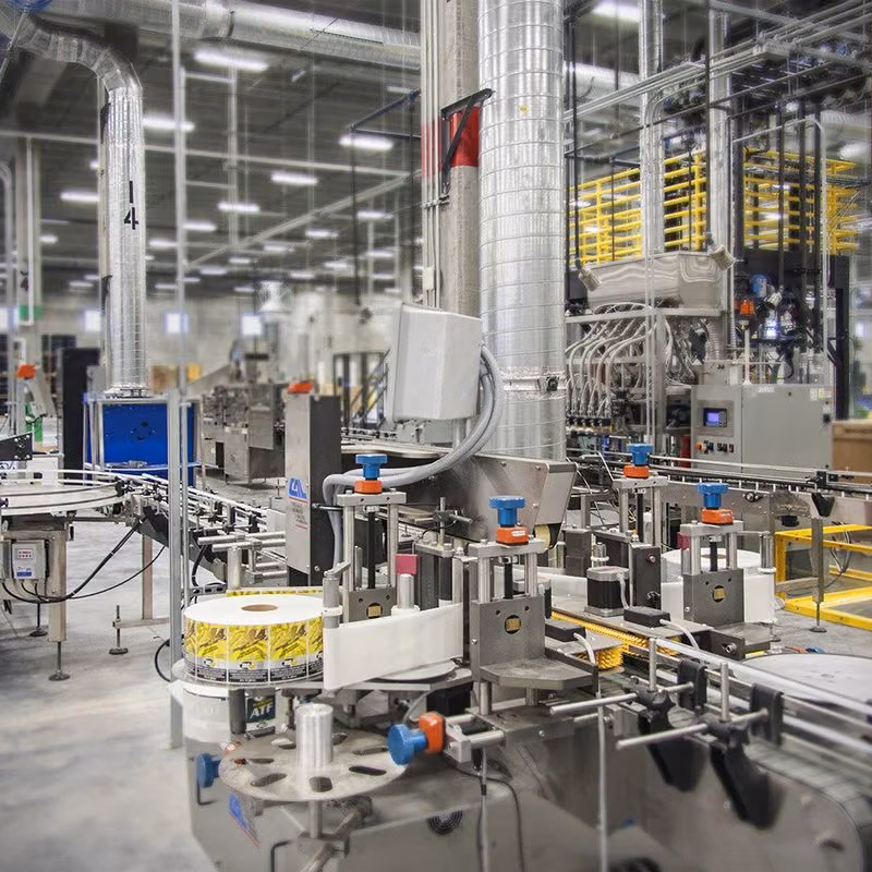 Stainless steel labeling machines on a factory production line, with cans or bottles moving along a conveyor.