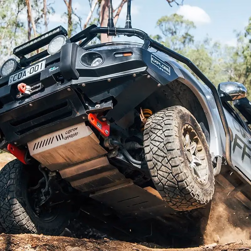 A silver off-road pickup climbing a dirt trail, showing its black bumper and a Custom Offroad skid plate under the body.