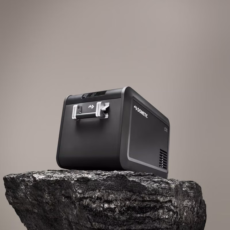 Black Dometic portable cooler with silver handle sitting on a dark rock against a grey background.