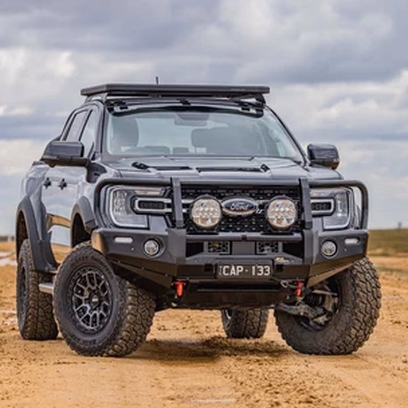 Black Ford pickup truck with a roof rack, bull bar and large offroad tyres parked on sand.