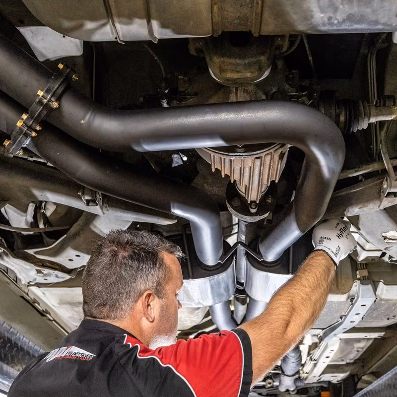 A mechanic in a black and red shirt works on the exhaust pipes underneath a car on a lift.