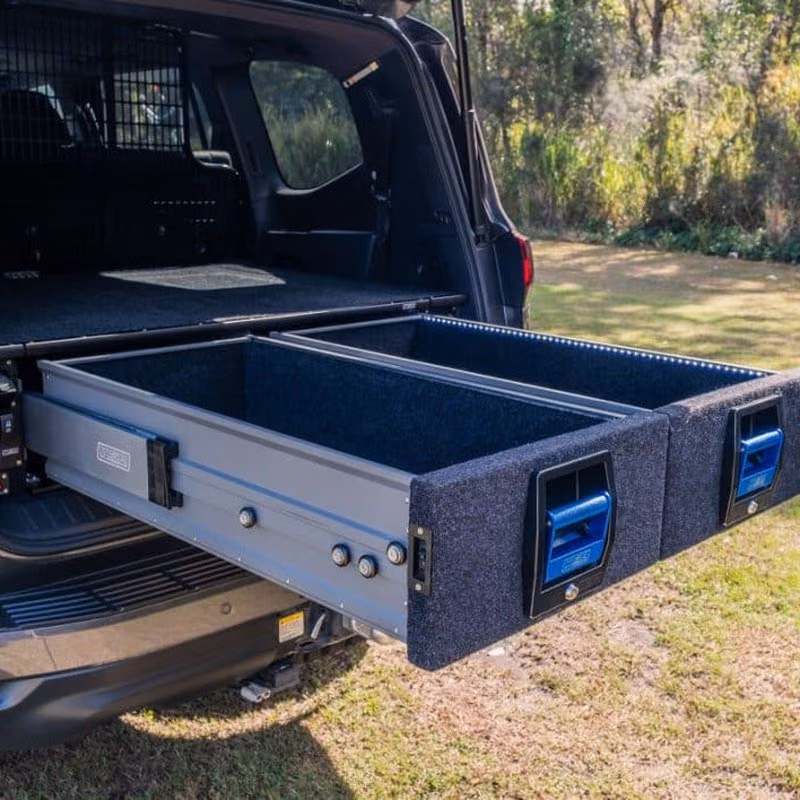 Two carpeted slide-out drawers with blue handles pulled out from the back of an SUV onto grass.