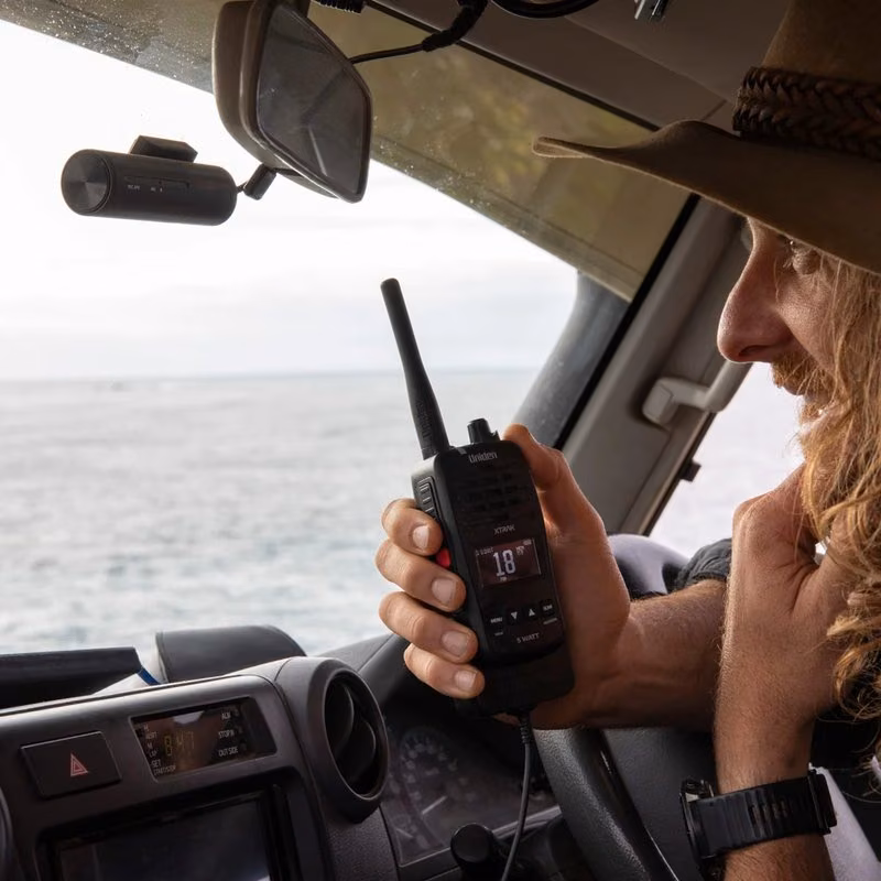 A man in a wide-brimmed hat holds a black Uniden handheld radio while driving, with the ocean visible through the windshield.